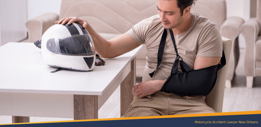 Sad man due to the motorcycle accident sitting on the chair with an arm sling on his left hand and resting his other hand on top of the motorcycle helmet