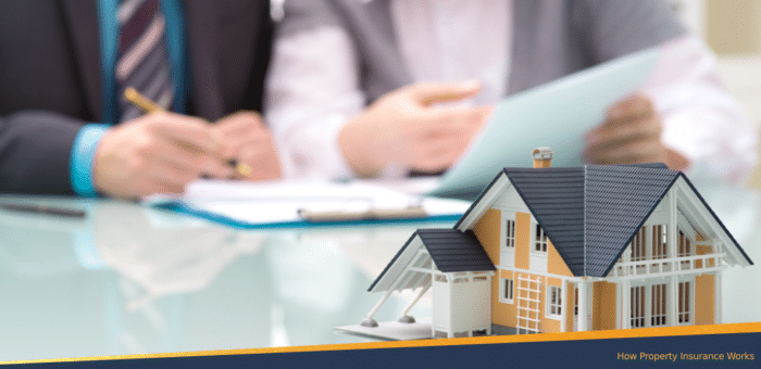 Miniature house model on a glass table with two people reviewing documents in the background, representing a discussion about property insurance coverage.