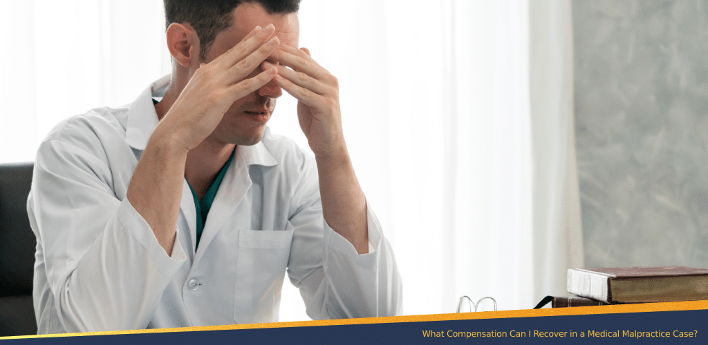 Stressed male doctor in a white coat sitting at a desk with hands on his face, symbolizing emotional strain often tied to medical malpractice cases.